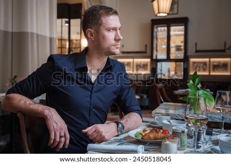 A man in a black shirt sits at a restaurant table with a croissant, red sauce, white wine, and a plant centerpiece. Warm ambiance with framed pictures. Location unknown.
