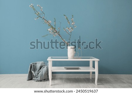 Vase with blooming branches, candles and laptop on glass coffee table near blue wall