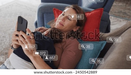 Image of social media icons with numbers over caucasian woman holding baby and using smartphone. Social media, communication and digital interface concept digitally generated image.