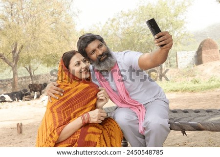 Young happy rural indian couple wearing traditional outfit sitting on bed taking selfie picture with smartphone .