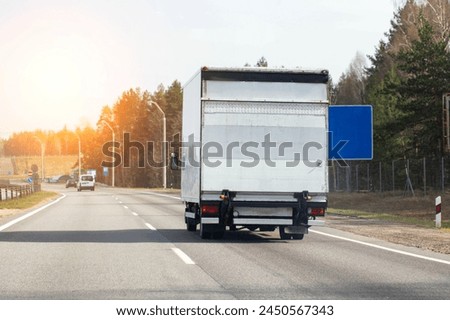 A modern van with a tail lift is driving along an asphalt road against the backdrop of a sunset. Concept of road transport of trucks with hydraulic lift. Copy space for text, bearing capacity Royalty-Free Stock Photo #2450567343