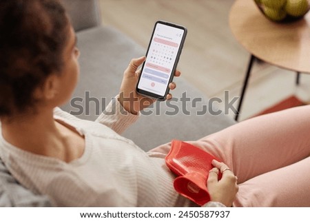 High angle closeup of young woman holding smartphone with period tracker app on screen and holding hot water bottle on belly copy space