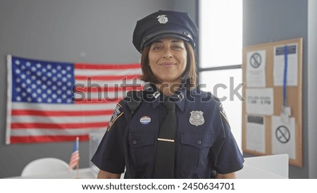 Smiling hispanic policewoman with badge, radio and 'i voted' sticker, in uniform, inside polling station with american flag. Royalty-Free Stock Photo #2450634701