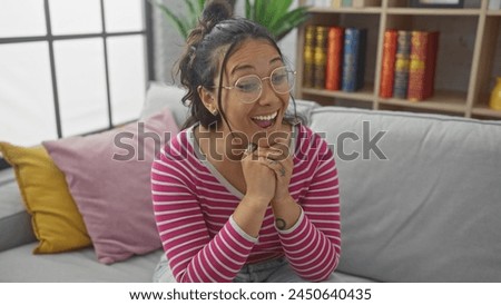 Excited young woman with glasses sitting on couch in brightly lit living room, expressing joy. Royalty-Free Stock Photo #2450640435