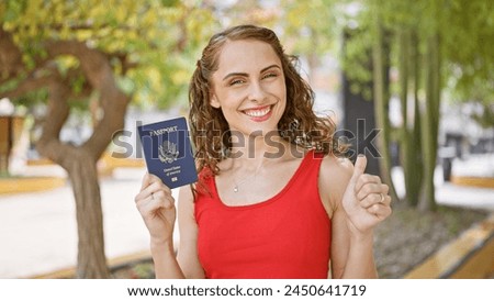 Radiant young woman, grinning ear-to-ear, holds up her american passport in the sunlight at the park, giving the thumb up gesture for her exciting upcoming travel adventure.