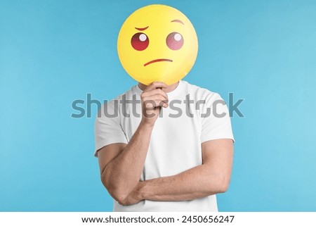 Man covering face with thinking emoticon on light blue background