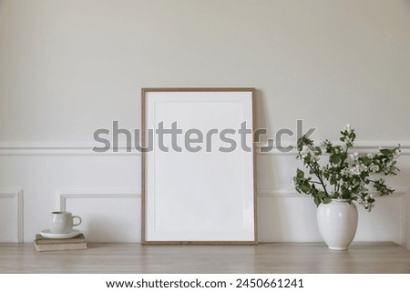 Springtime breakfast still life. Empty picture frame poster mockup. White ceramic vase with blooming apple tree branches. Cup of coffee, tea on wooden table, desk with old books, scandi home interior