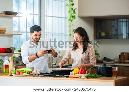 Indian young couple cooking food in kitchen and taking selfie picture using smartphone or filming recipe