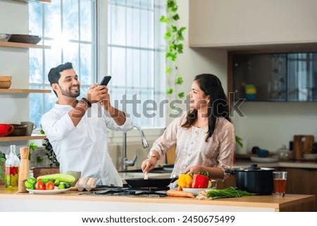 Indian young couple cooking food in kitchen and taking selfie picture using smartphone or filming recipe
