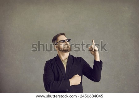 Funny guy thinks of a very smart idea. Young man in a suit jacket and thug life 8 bit meme glasses pointing his finger up isolated on a grey background