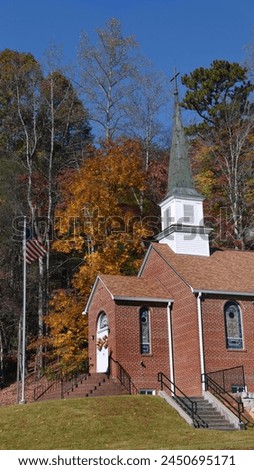 The Mt. Hebron Wesleyan Church is surrounded by Autumn color.  Exterior of building is red brick and it has a steeple topped by a cross. Royalty-Free Stock Photo #2450695171