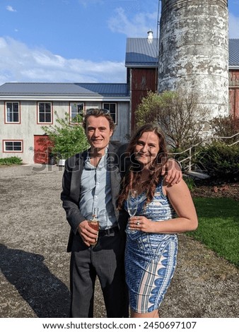 A well-dressed couple holding drinks take a picture outside at a barn wedding