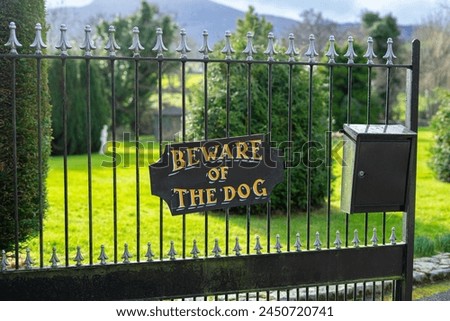 Hand painted "beware of the dog" sign in yellow artsy font on black big plaque hung on metal steal gate entrance fence of wealthy home letterbox bright green grass and trees on front lawn warning
