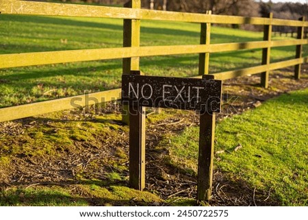 Wooden national park sign posted that says NO EXIT in white letters on wood plank in ground in front of wooden fence grassy area early morning sunlight to control traffic dead end no car entrance