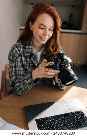Vertical portrait of young female photographer holding modern digital camera looking at screen choosing photos for editing while sitting in front of laptop computer at home office workplace.
