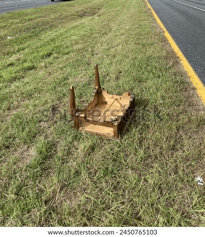 A small wooden end table is on the grass median of a black-surfaced road. It is out of place, upside-down, and missing two legs. Perhaps the tables are turned.  Royalty-Free Stock Photo #2450765103