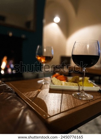 An inviting evening scene with glasses of red and white wine on a wooden table, a cozy fireplace in the background providing a gentle glow, and a platter of cheese and fruit completing this picture of