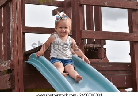 Little blonde haired girl with a bow in her hair, sitting at top of slide, playground, summer time. Royalty-Free Stock Photo #2450780091