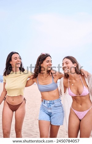 Vertical pic of Three young Caucasian beautiful women friends embracing in swimwear posing cheerful on beach holidays. Group of gen z female hugging standing smiling on sunny summer time