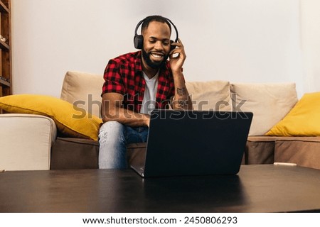 Black man in casual clothes and headphones sitting on the sofa using laptop. The image conveys the concept of watching movies and videos on the computer, but also teleworking and video calls at home.