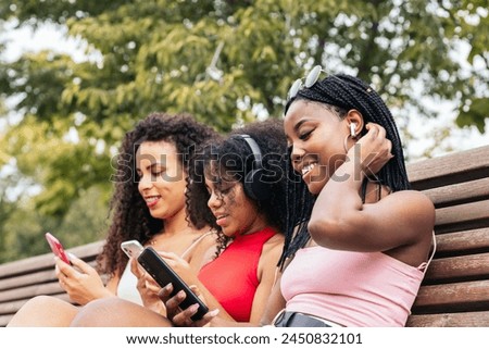 African american female friends sitting on a park bench laughing and having fun while using smartphone and listening to music with headphones Royalty-Free Stock Photo #2450832101