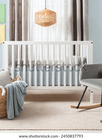 Serenely styled baby's bedroom highlighting a white modern crib, a plush gray rocker and a beige pattern rug. The soft daylight filters through sheer curtains beside a woven rattan pendant light. Royalty-Free Stock Photo #2450837793