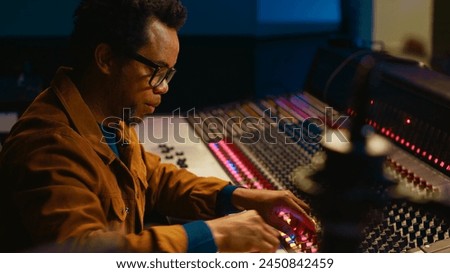 Music producer using audio mixer in professional studio, adjusting volume levels while pressing buttons and sliders on control desk. Sound designer works with digital audio software. Camera A.