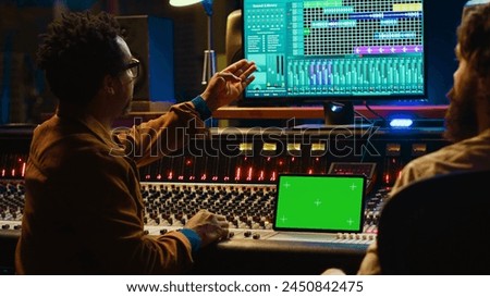 Professional engineer and musician mixing tracks next to isolated display on tablet, editing audio after recording session to produce a hit song. Artist collaborates with sound designer. Camera A.