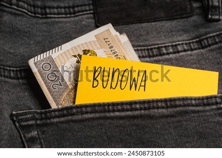 Yellow card with a handwritten inscription "budowa", inserted into the pocket of gray pants jeasnow, next to Polish banknotes PLN (selective focus), translation: construction