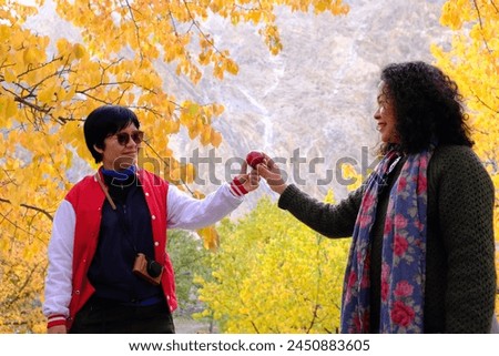 Two Asian female sisters holding an apple, posing for a picture with a view of the colorful foliage in the morning light in Passu Valley, Gilgit-Baltistan, Pakistan.