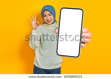 Cheerful beautiful Asian woman in white sweater and hijab, showing blank mobile phone screen, showing okay gesture isolated on yellow background. Muslim lifestyle concept