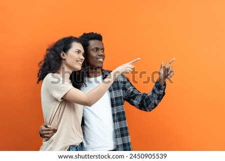 Happy Couple Pointing On Orange Background, Young People, Diversity, Smiling, Casual Clothing, Togetherness, Positive Emotion, Lifestyle Concept, African American, Mixed Ethnicity, Relationship Goals. Royalty-Free Stock Photo #2450905539