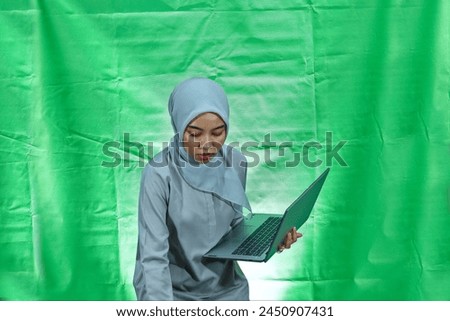 beautiful young Asian woman wearing hijab and blouse holding laptop and taking someting below isolated on green background