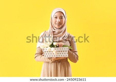 Beautiful young Asian woman in hijab with gift box and flowers on yellow background
