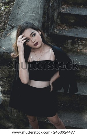 An attractive young Asian woman displays a stylish goth-inspired outfit while thoughtfully posing beside weather-worn steps in a serene setting.