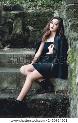 A beautiful young Asian woman dressed in goth-inspired attire poses elegantly by weathered stone steps, emanating a mystical and stylish aura.