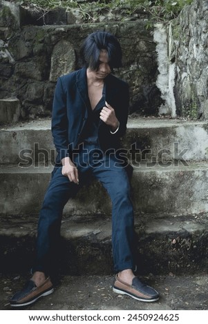 A young Asian man exhibits a unique style with goth-inspired clothing sitting on concrete steps, set against an atmospheric damp backdrop.