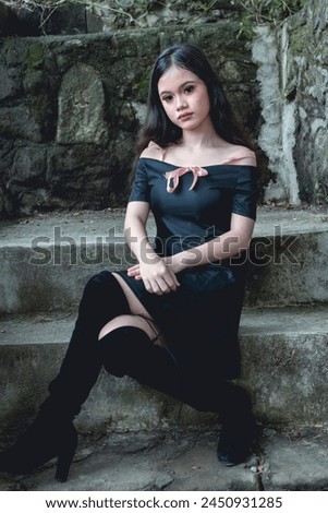 A young woman dressed in goth-inspired fashion sits elegantly on stone steps, elegantly posing against weathered and damp concrete steps