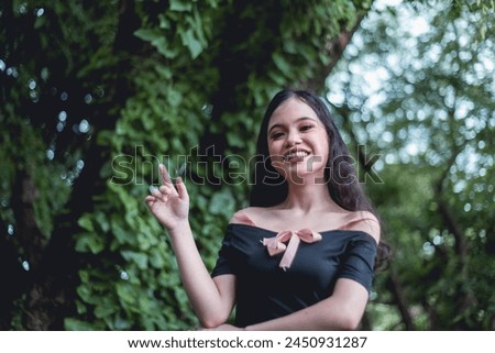 Captured in a serene woodland setting, a young woman sports goth-inspired clothing while smiling broadly and gesturing a peace sign.