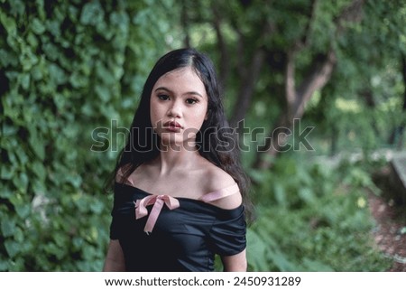 Enigmatic young woman wearing goth-inspired outfit stands amidst a verdant forest, enhancing the mysterious and ethereal atmosphere.