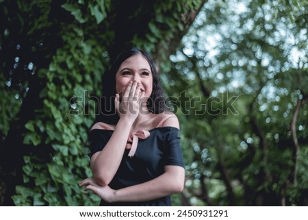 A captivating young woman in a goth-inspired black dress with a pink ribbon, laughing joyfully in a serene forest background.