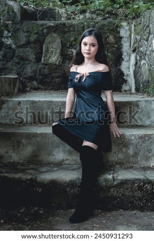 A young woman wears a goth-inspired dress and knee-high boots, elegantly posing against weathered and damp concrete steps
