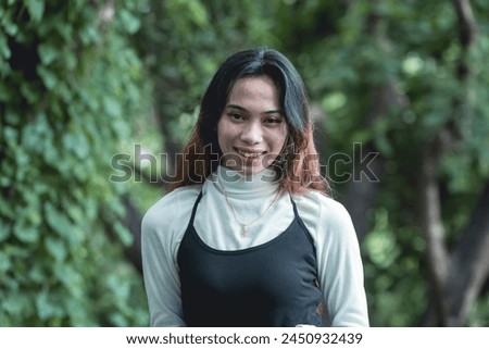 A portrait of a young woman dressed in goth-inspired clothing, smiling gently with a natural forest as her background.