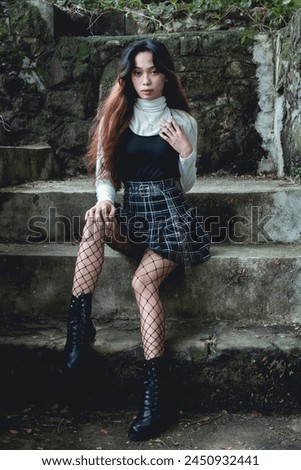 A young woman in a goth-inspired outfit sits confidently on stone steps in a forest, exuding a mysterious aura.