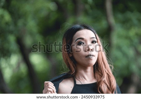 A portrait of a young woman dressed in goth fashion, set against a vibrant green forest background, embodying a sense of bold identity.