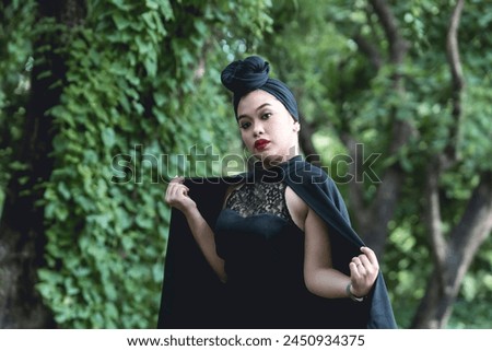 A stylish young woman clad in goth-inspired fashion poses confidently against a dense, green forest background, projecting a blend of elegance and mystery.