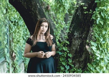 A young woman in a black goth-inspired dress stands thoughtfully in a forest, surrounded by green foliage and ancient trees.