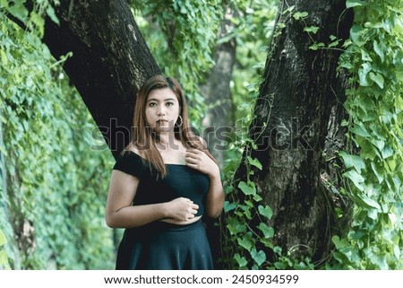 A young woman dressed in a stylish goth-inspired black outfit stands confidently in a verdant forest, exuding a mysterious aura.