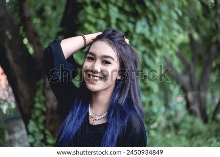 A cheerful young woman wearing goth-style attire poses with a bright smile in a verdant forest background, exuding happiness and confidence.