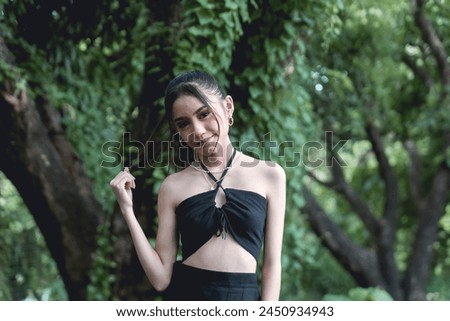 A stylish young woman dressed in a goth-inspired black outfit stands amid a dense, green forest, showcasing urban alternative fashion outdoors.